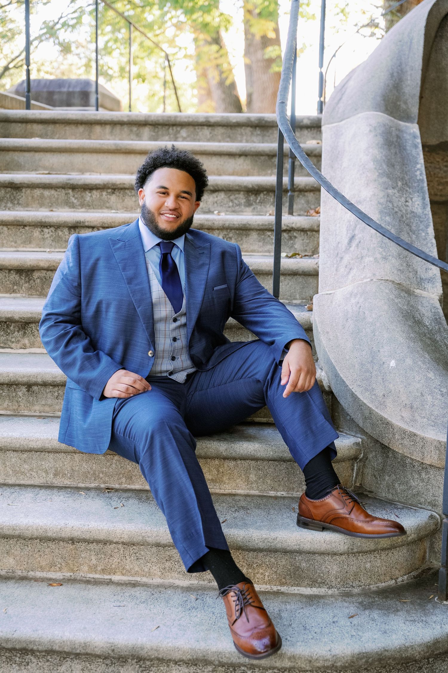 A person in a navy blue suit and vest sits casually on concrete steps while wearing brown leather dress shoes.
