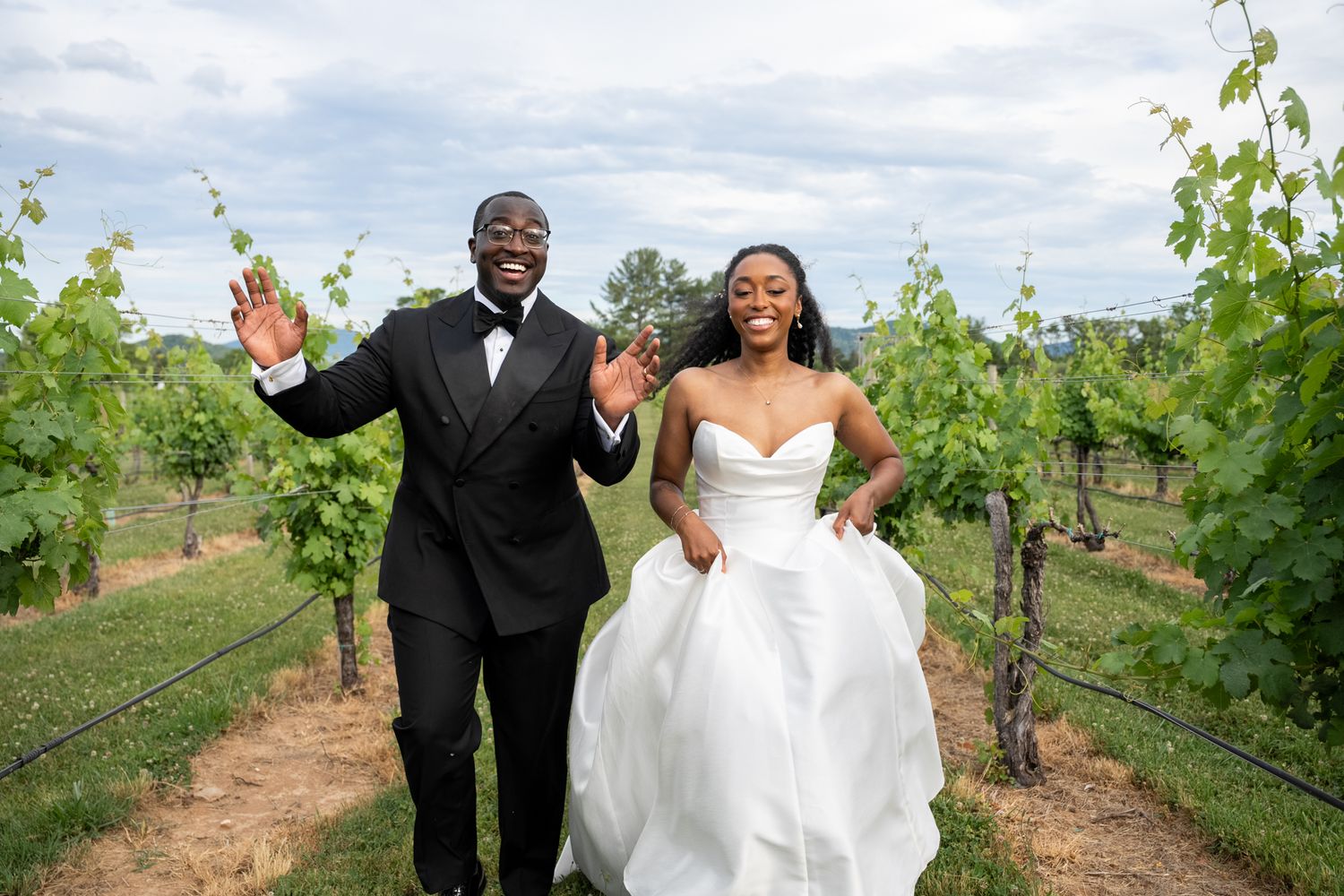Joyful newlyweds celebrate in a vineyard, bride in white gown and groom in black tuxedo, waving among green grapevines.