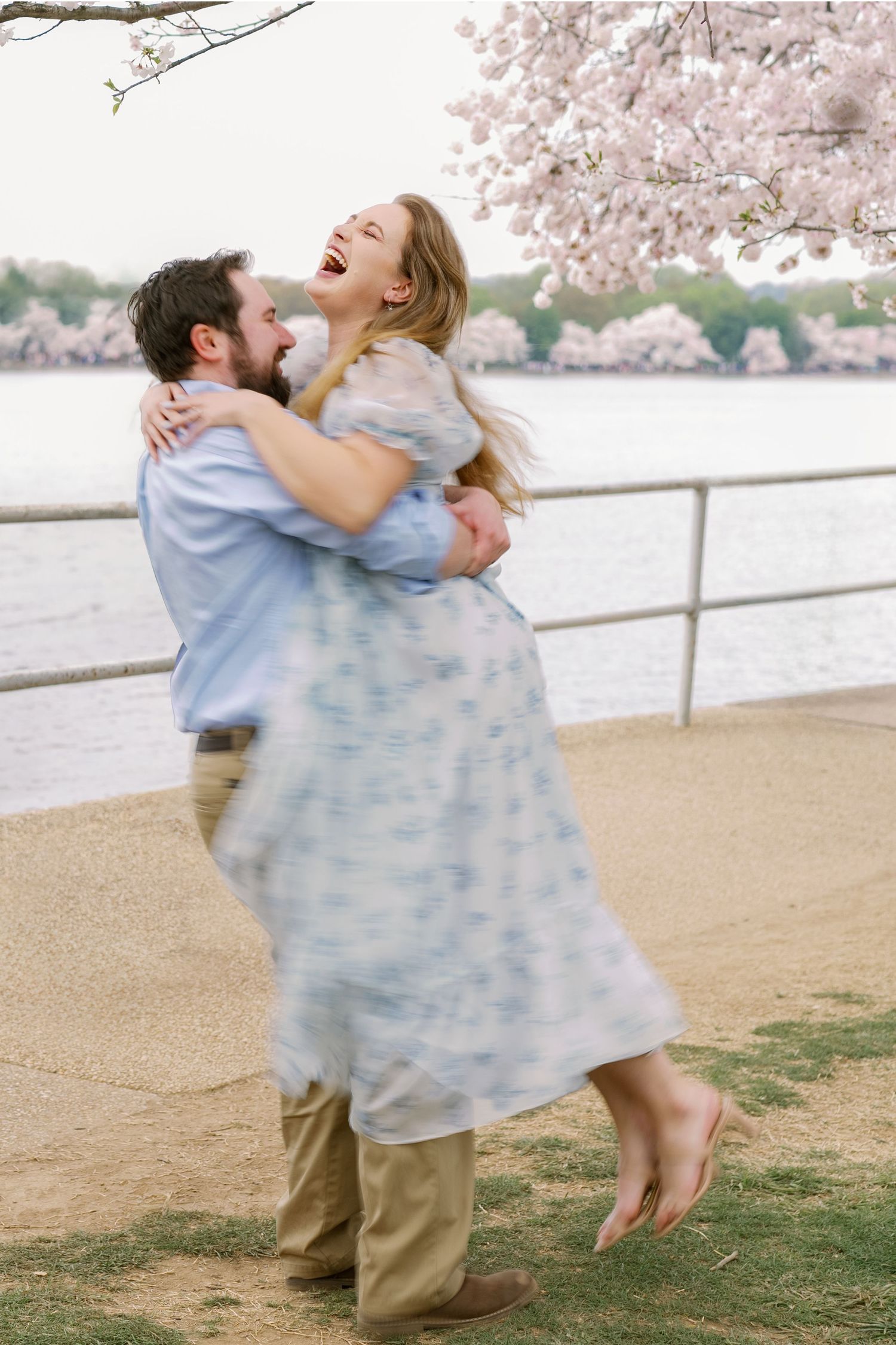Joyful couple embrace and laugh while standing by waterfront with cherry blossoms in background.