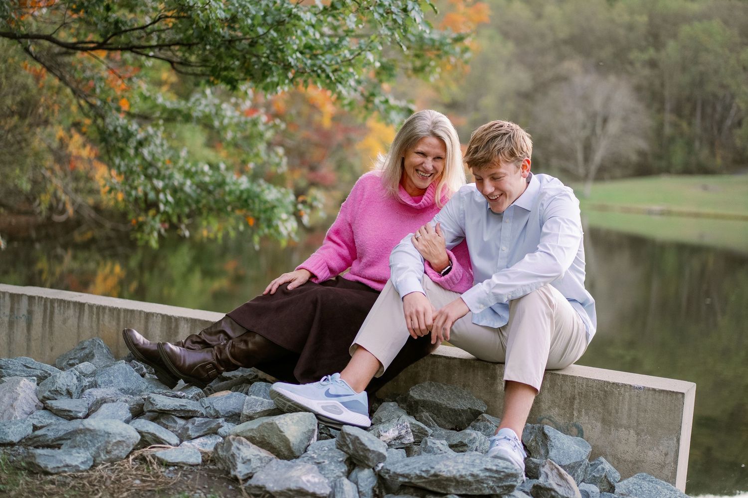 A couple sits together on rocks near a pond surrounded by autumn foliage during an outdoor photo session.