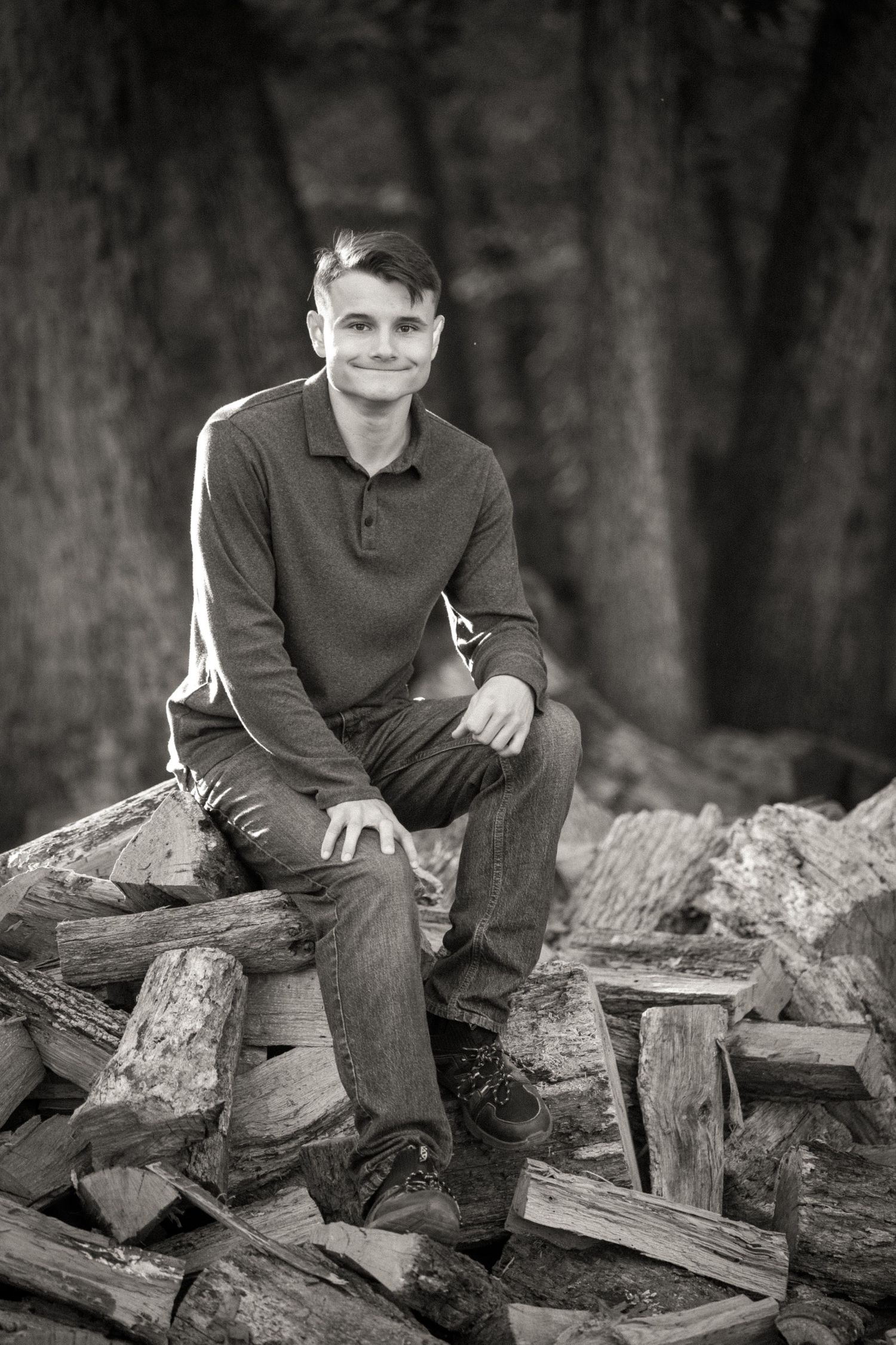 A black and white photo of someone sitting on a pile of firewood logs in a rustic outdoor setting.