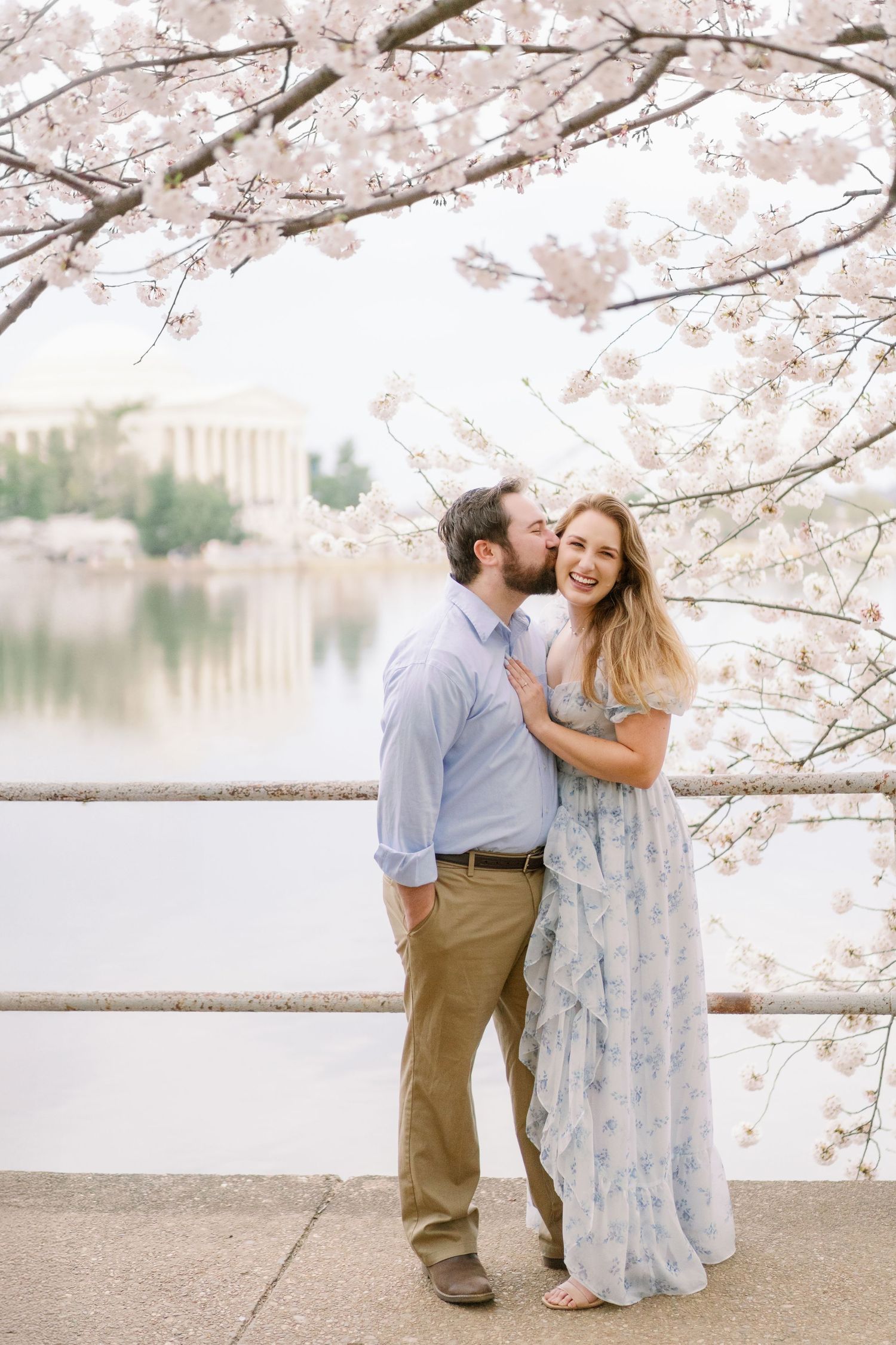 A couple embraces under cherry blossoms with the Jefferson Memorial visible in the background.