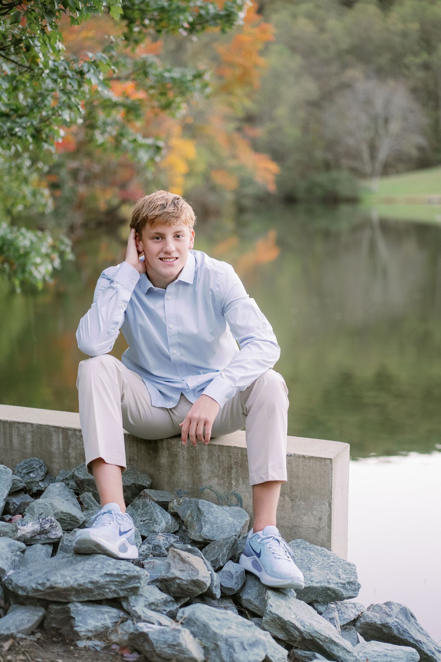 Outdoor portrait session on rocky waterfront with autumn foliage and pond in background.