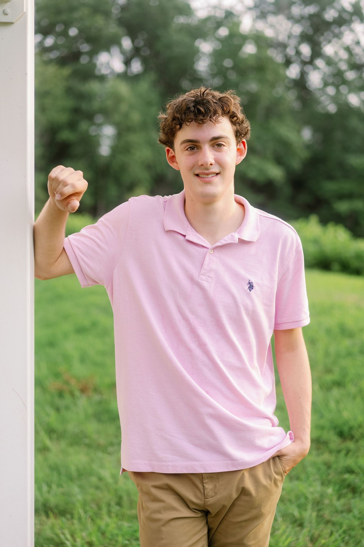 Young person in pink polo shirt and khaki pants poses outdoors against green foliage background.