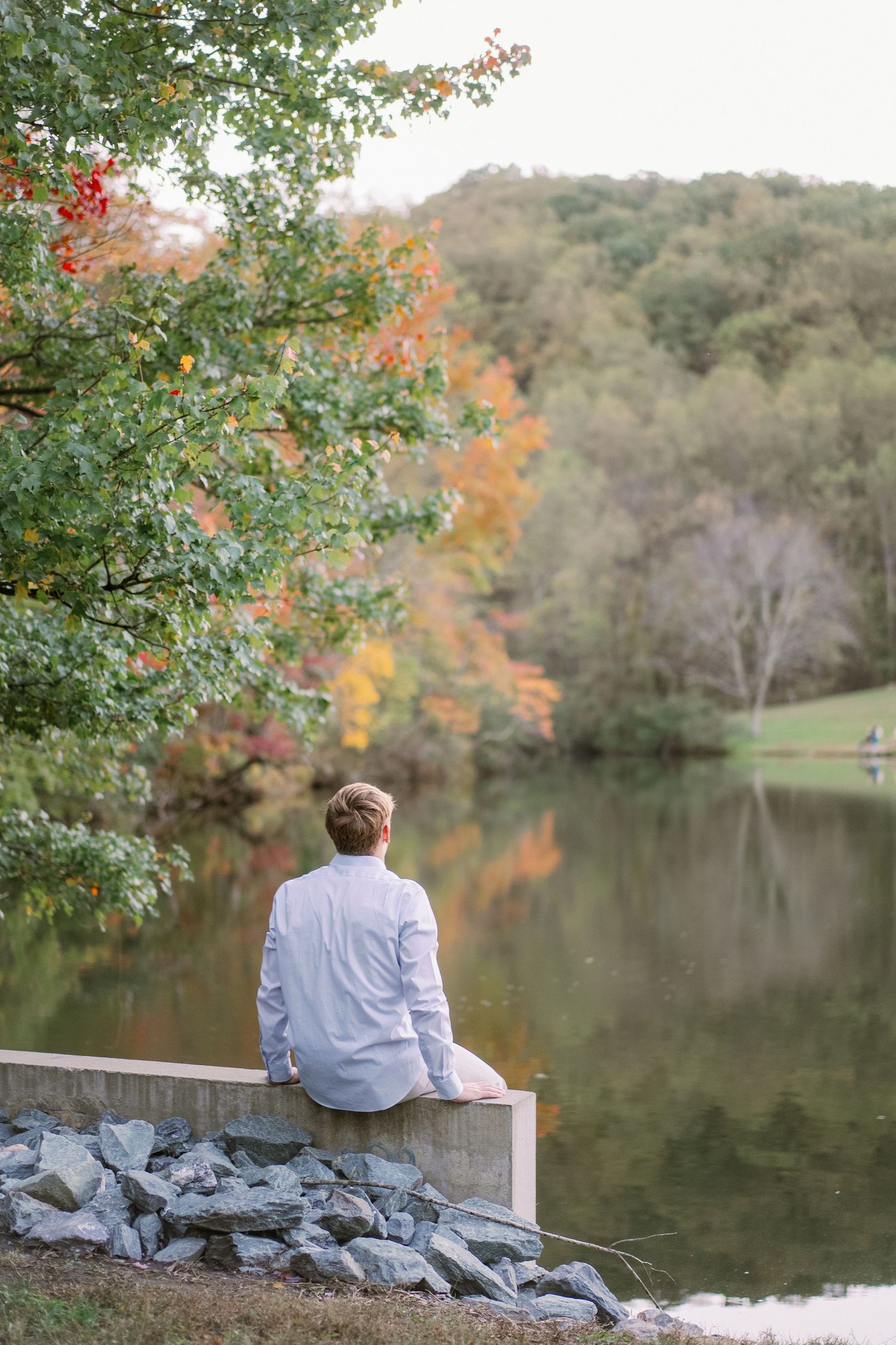A person in a white shirt sits peacefully on rocks by a serene lake surrounded by autumn foliage and trees.