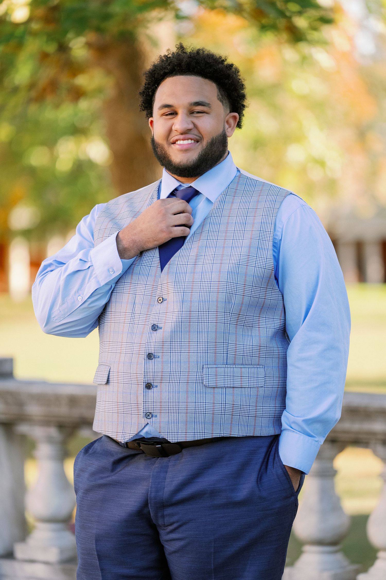 A person wearing a light blue dress shirt and plaid vest smiles while posing outdoors.