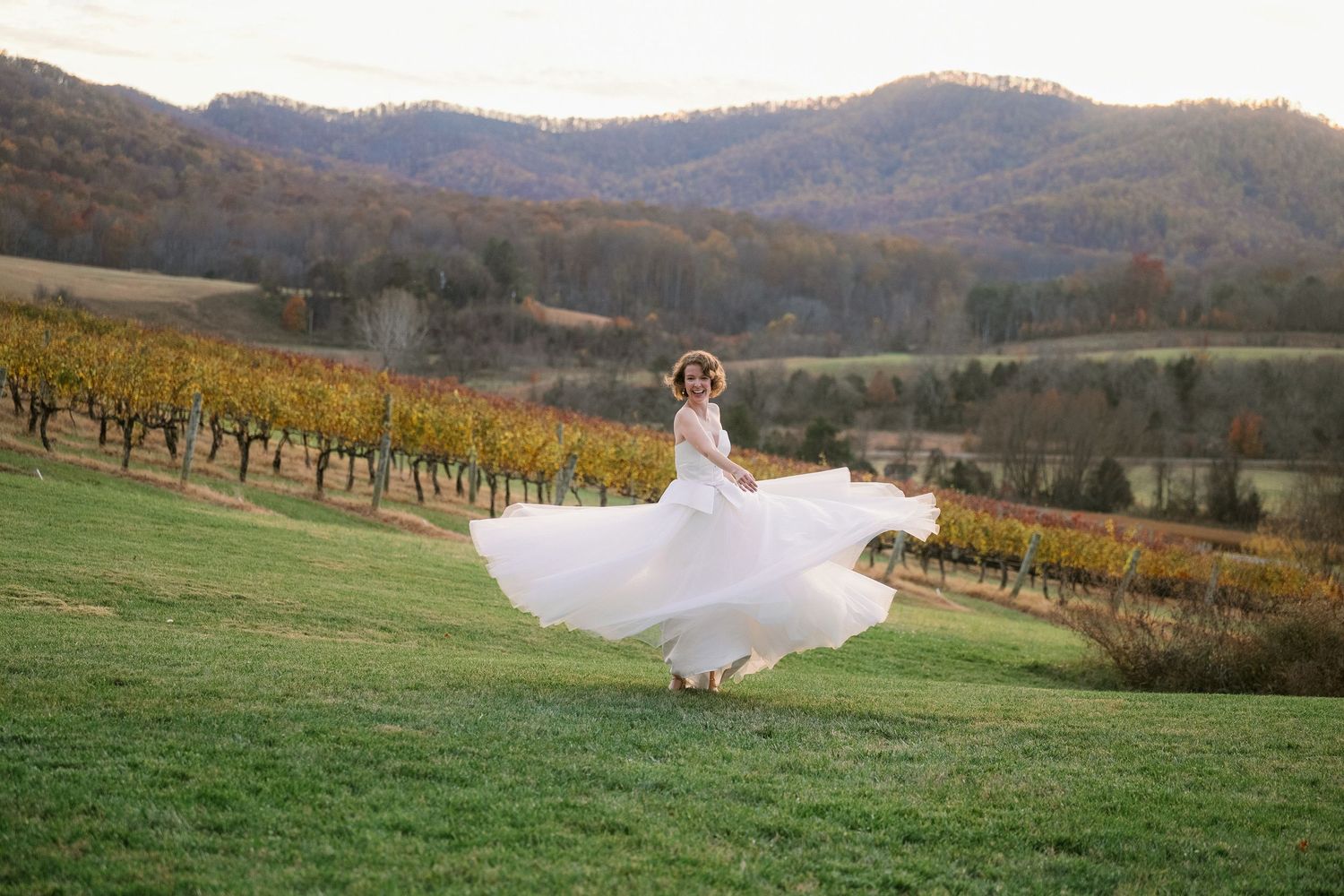 A woman in a flowing white dress twirls in a lush vineyard with rolling hills in the background.