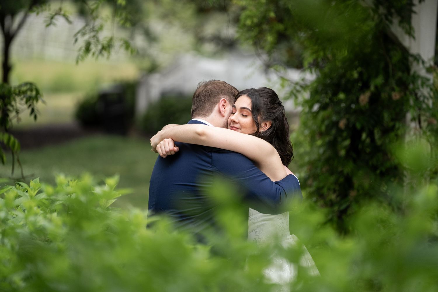 A bride and groom share a tender embrace surrounded by lush green foliage during their wedding day outdoor portrait.