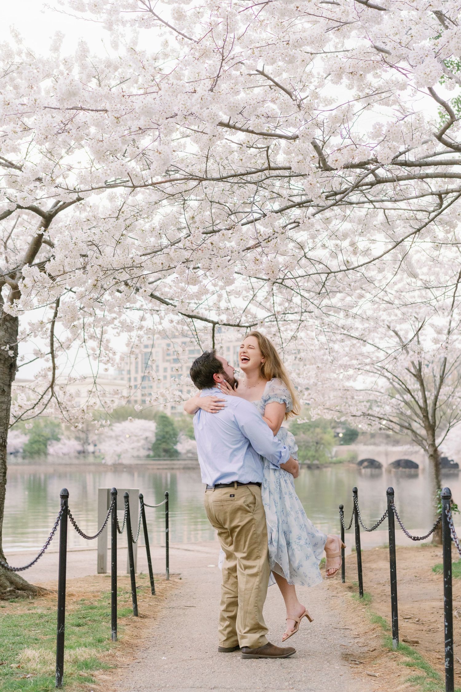 A joyful moment under blooming cherry blossoms along the Tidal Basin with a bridge in the background.