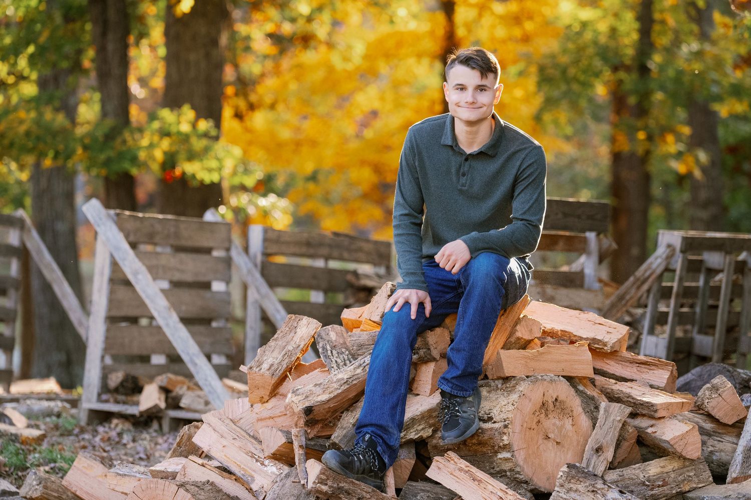 Child in blue sweater plays on rustic woodpile during autumn with golden foliage in background.