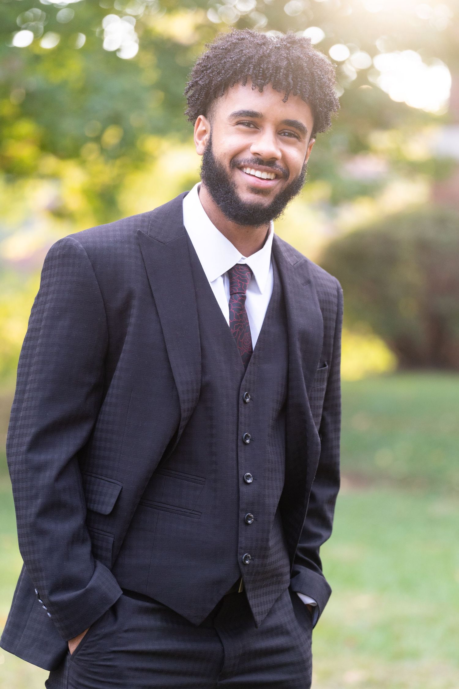 A person in a black three-piece suit poses confidently against a sunlit natural background.