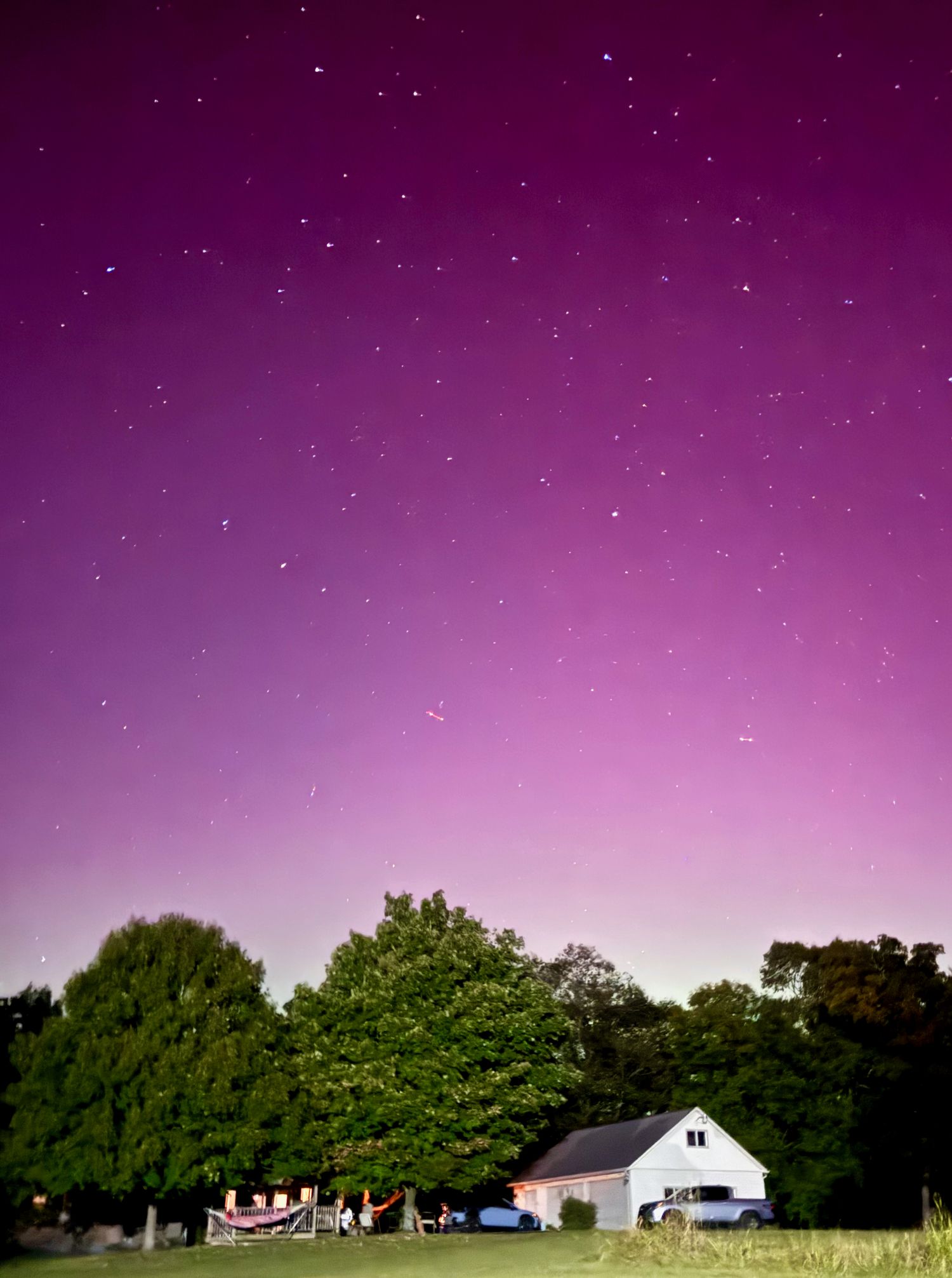 Purple aurora borealis illuminates the night sky above a white barn and trees in a rural field.