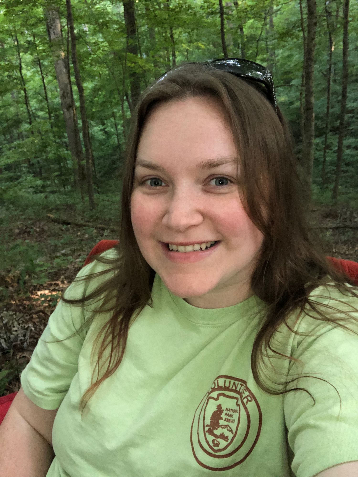 A person wearing a light green t-shirt with an emblem smiles while sitting in a lush forest setting.