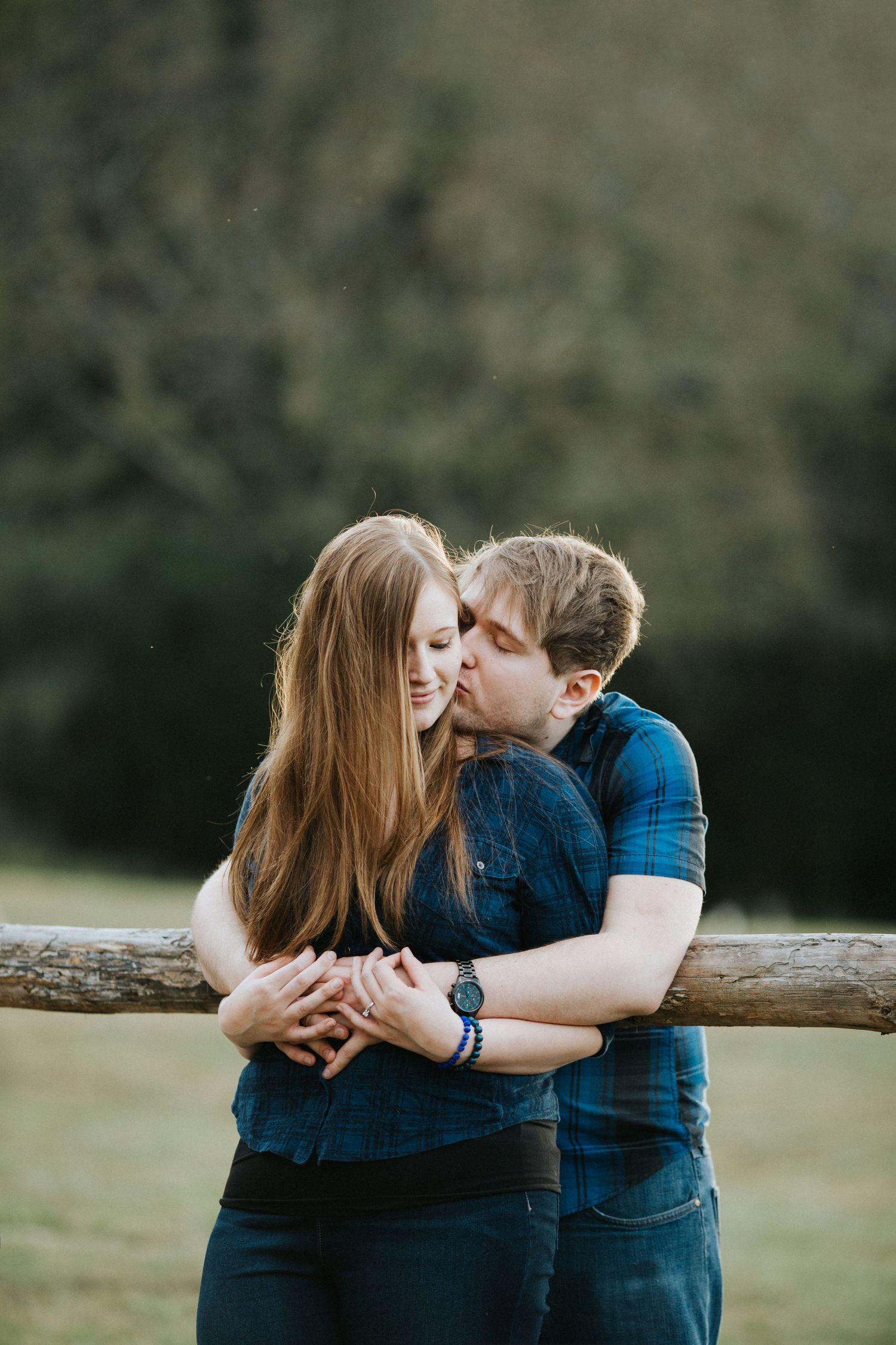 A couple embraces near a wooden fence outdoors, the man kissing the woman's cheek as she smiles warmly.