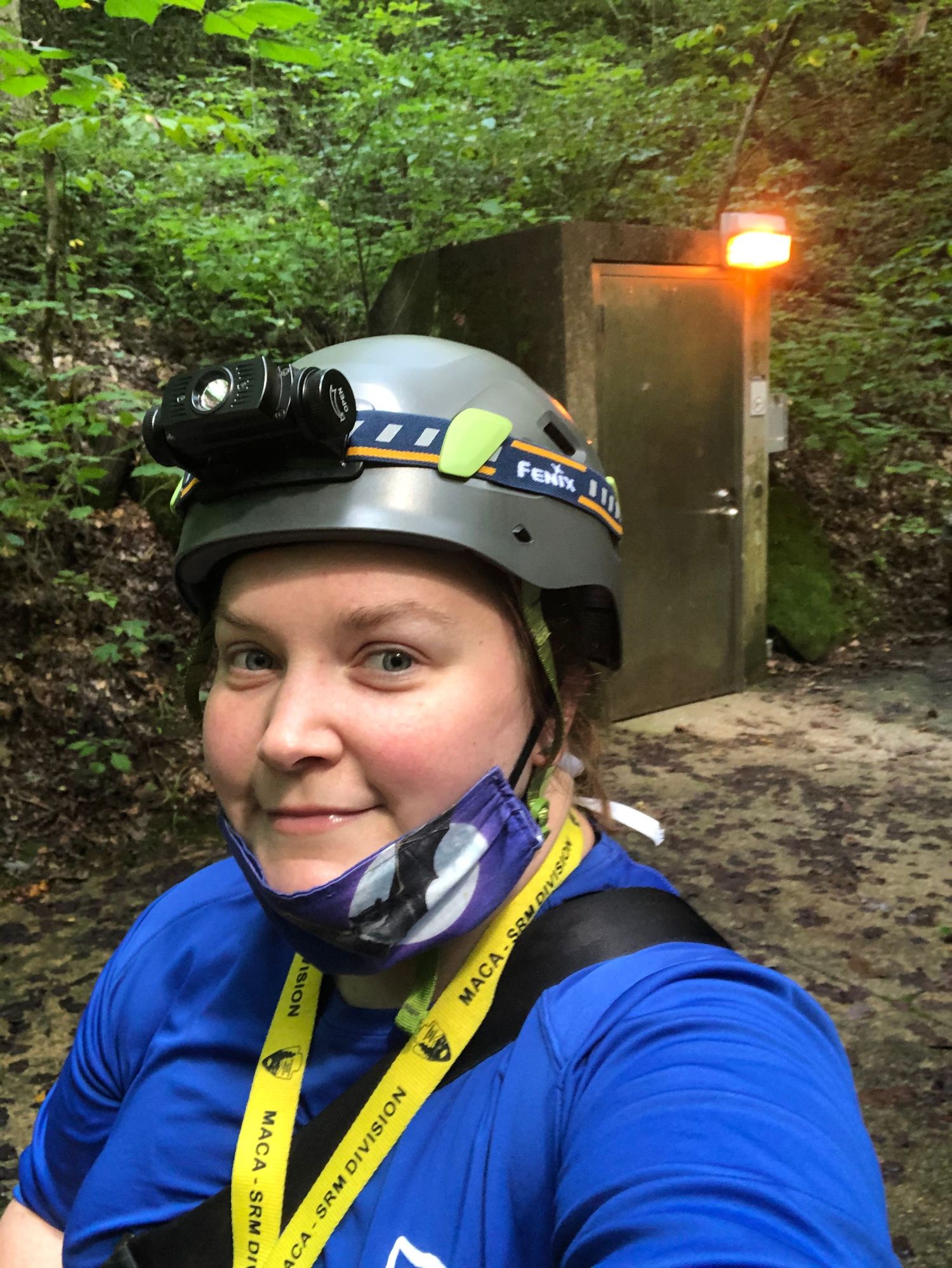 A person wearing a safety helmet and blue shirt stands at the entrance of an illuminated tunnel or bunker in a forested area.