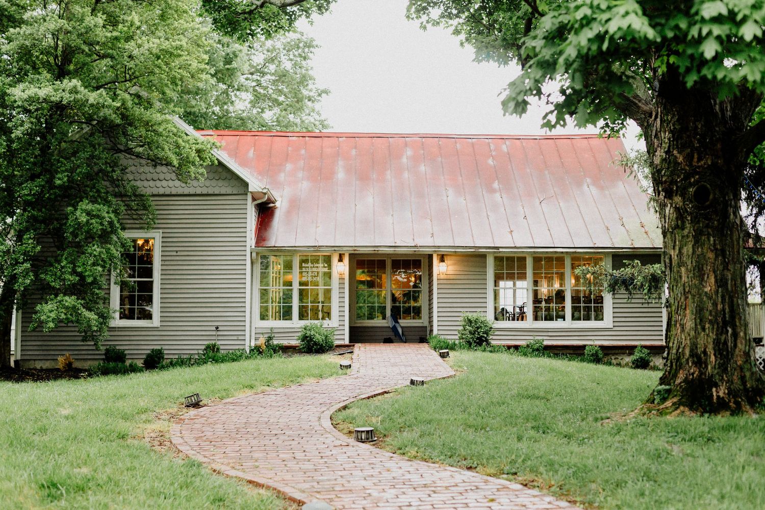 A curved brick pathway leads through a green lawn to a gray house with a red metal roof and white trim windows.