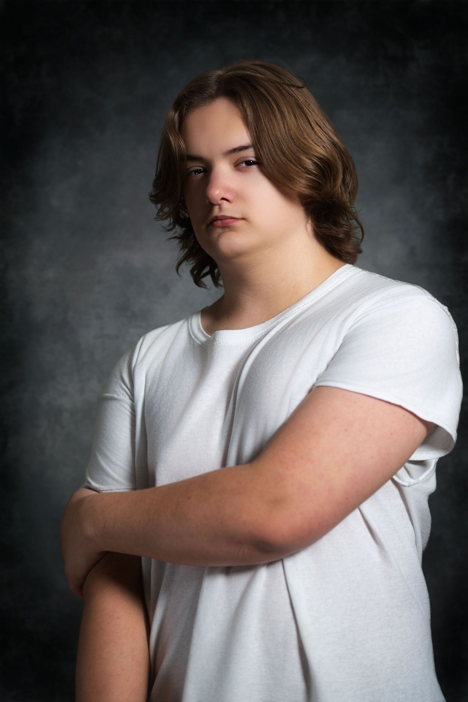 A teenage boy with shoulder-length brown hair wearing a white t-shirt poses with arms crossed against a dark studio background.