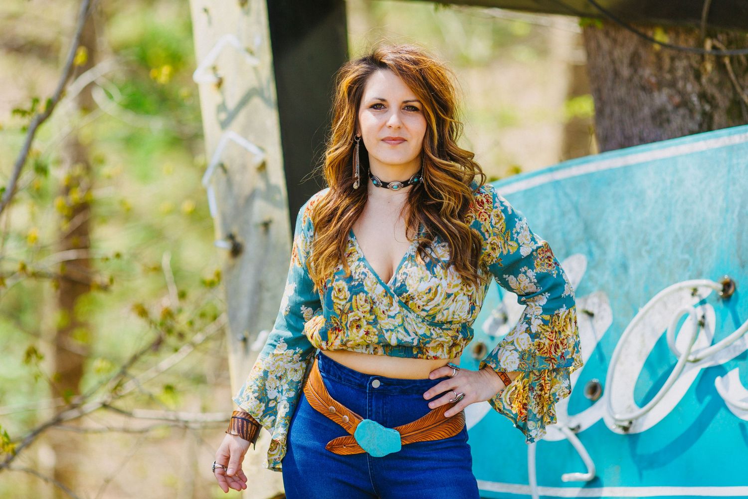 Woman in floral crop top and high-waist jeans poses outdoors near vintage turquoise sign.