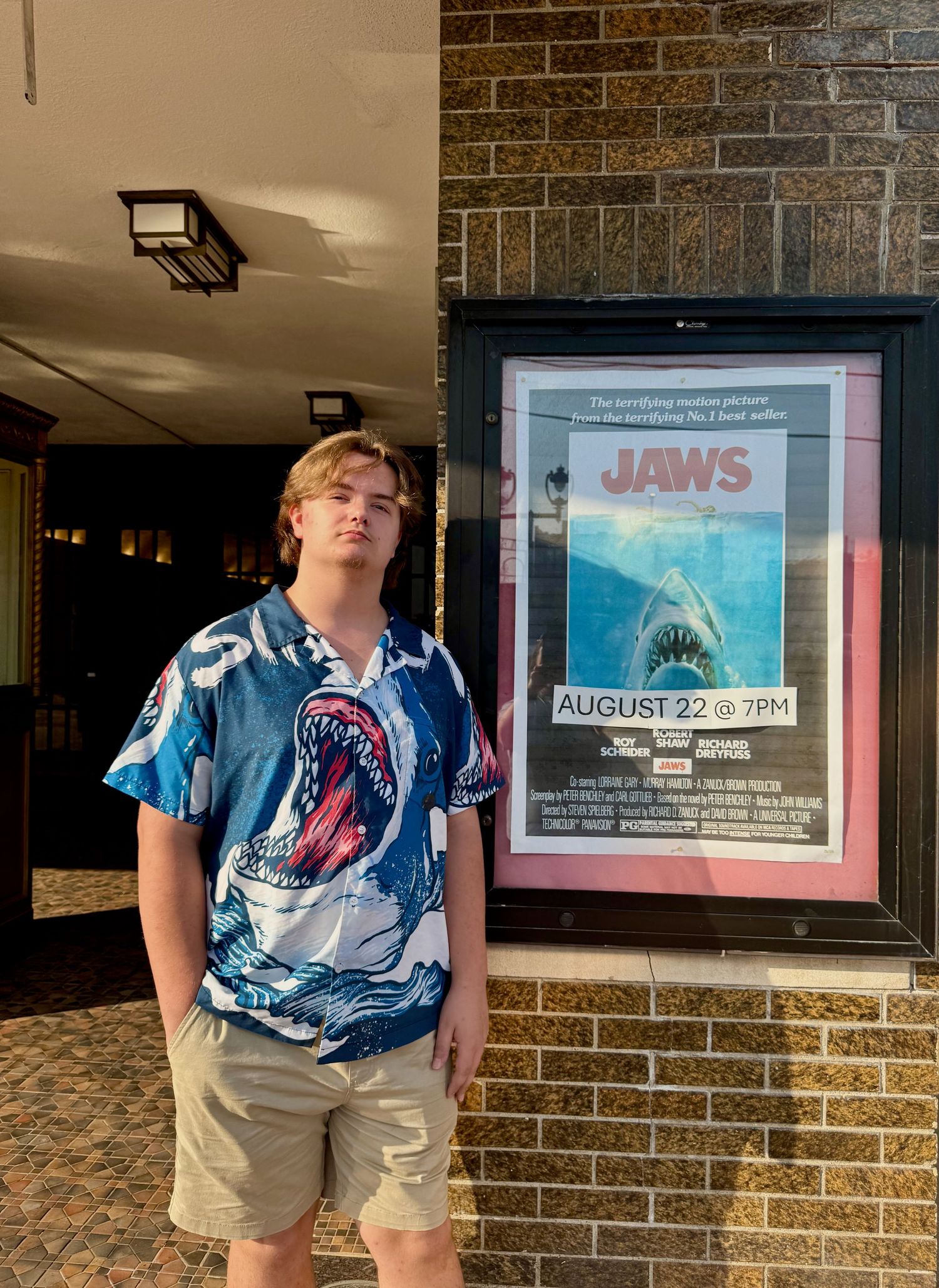 Young man in shark-print shirt stands beside a Jaws movie poster advertising an August 22 7PM screening outside a theater.