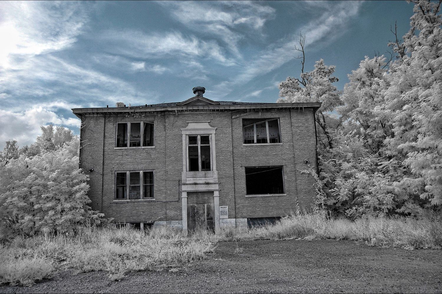 An abandoned two-story brick house with broken windows stands alone against a dramatic sky and white foliage.