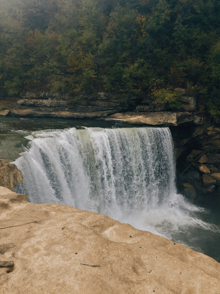 A powerful waterfall cascades over rocky ledges into a serene river, surrounded by lush green forest in a natural gorge.