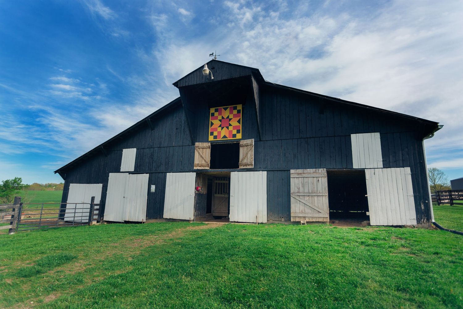 Black barn with colorful quilt square art on facade, white doors, green grass foreground, and blue sky with clouds.