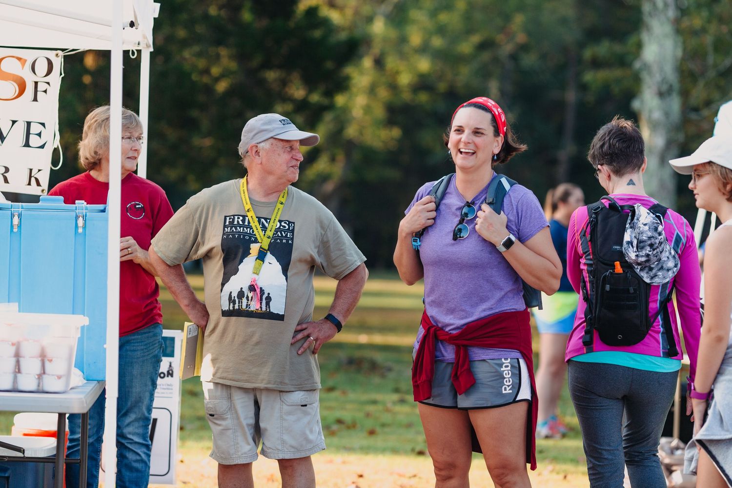 Smiling woman in red headband and backpack talks with older man wearing a medal at an outdoor running event.