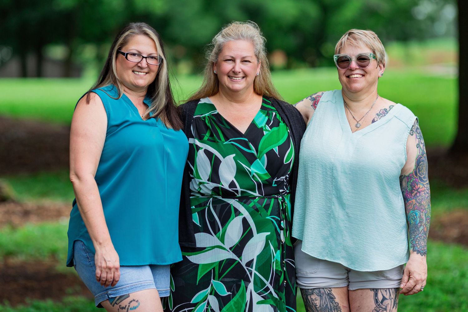 A group wearing coordinated green and teal outfits poses together outdoors in a park setting.