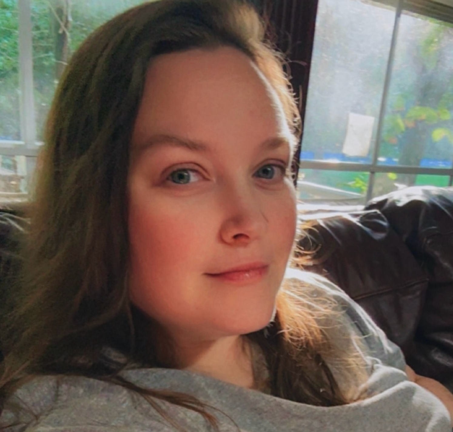 Young woman with long brown hair and blue eyes relaxing indoors near a window with natural light.