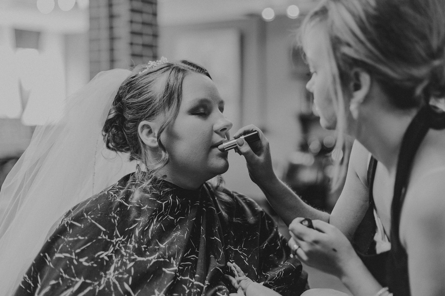 Bride getting lipstick applied by makeup artist before wedding ceremony, wearing veil and cape.