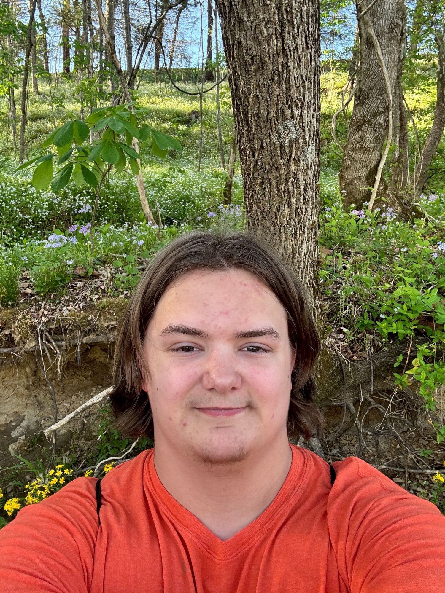 Young person in orange shirt takes selfie in lush spring forest with wildflowers and trees in background.