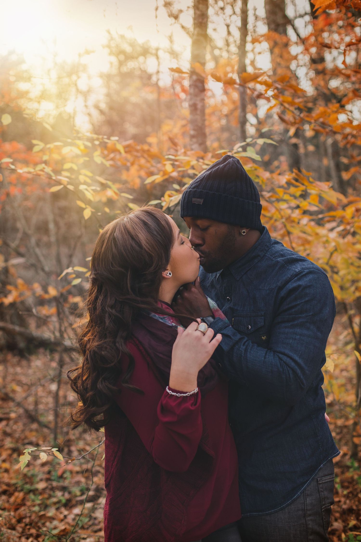Interracial couple sharing a romantic kiss in autumn forest with golden sunlight and colorful fall foliage backdrop.