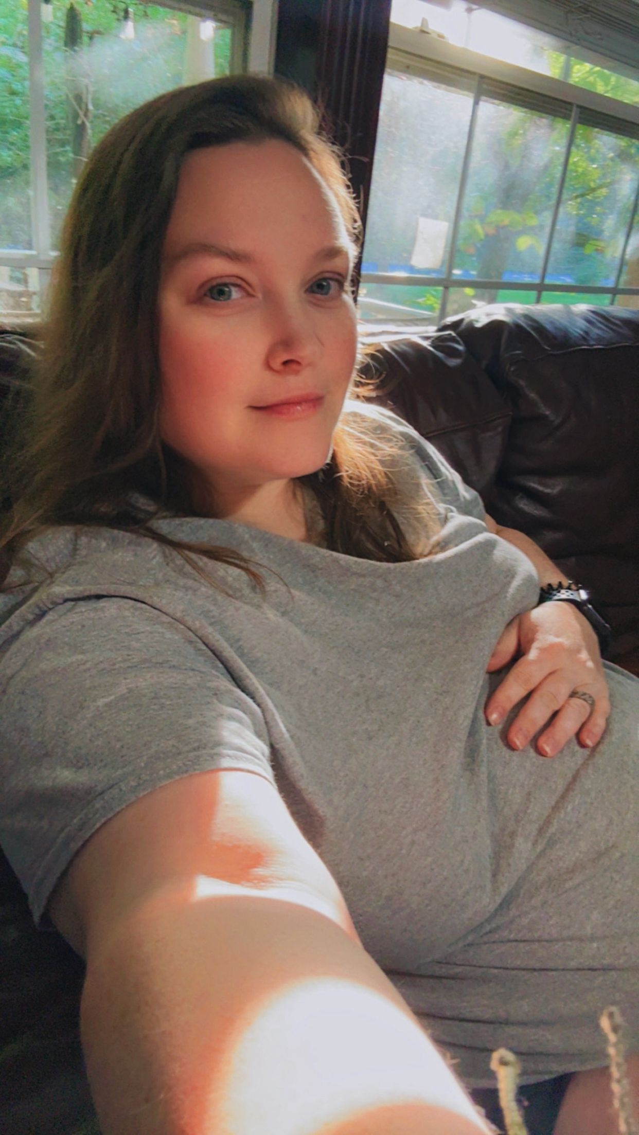Young woman with brown hair taking a selfie on a leather couch, wearing a gray t-shirt in a sunlit room.