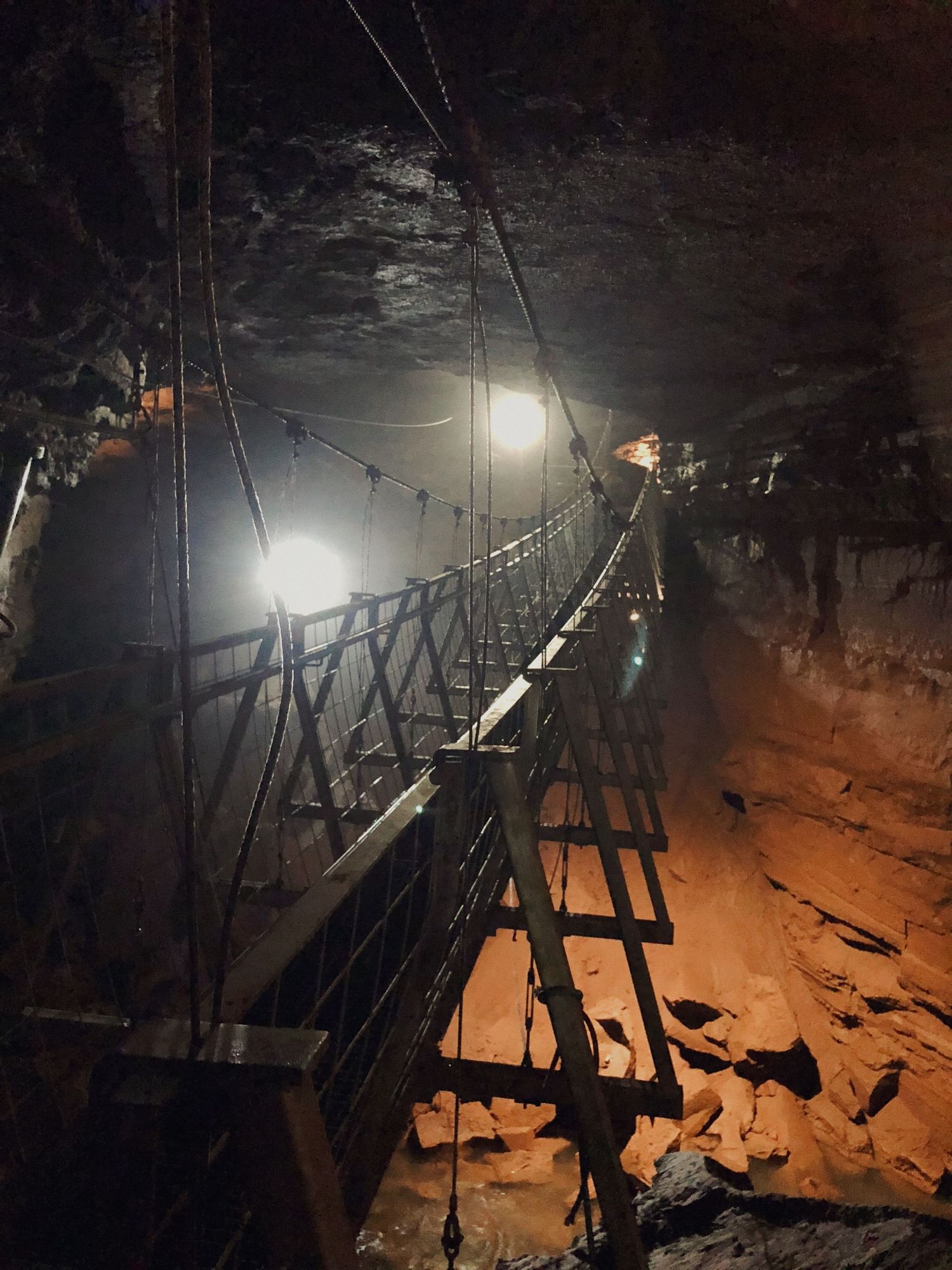 A suspension bridge illuminated by lights inside a dark cave with red rock formations.