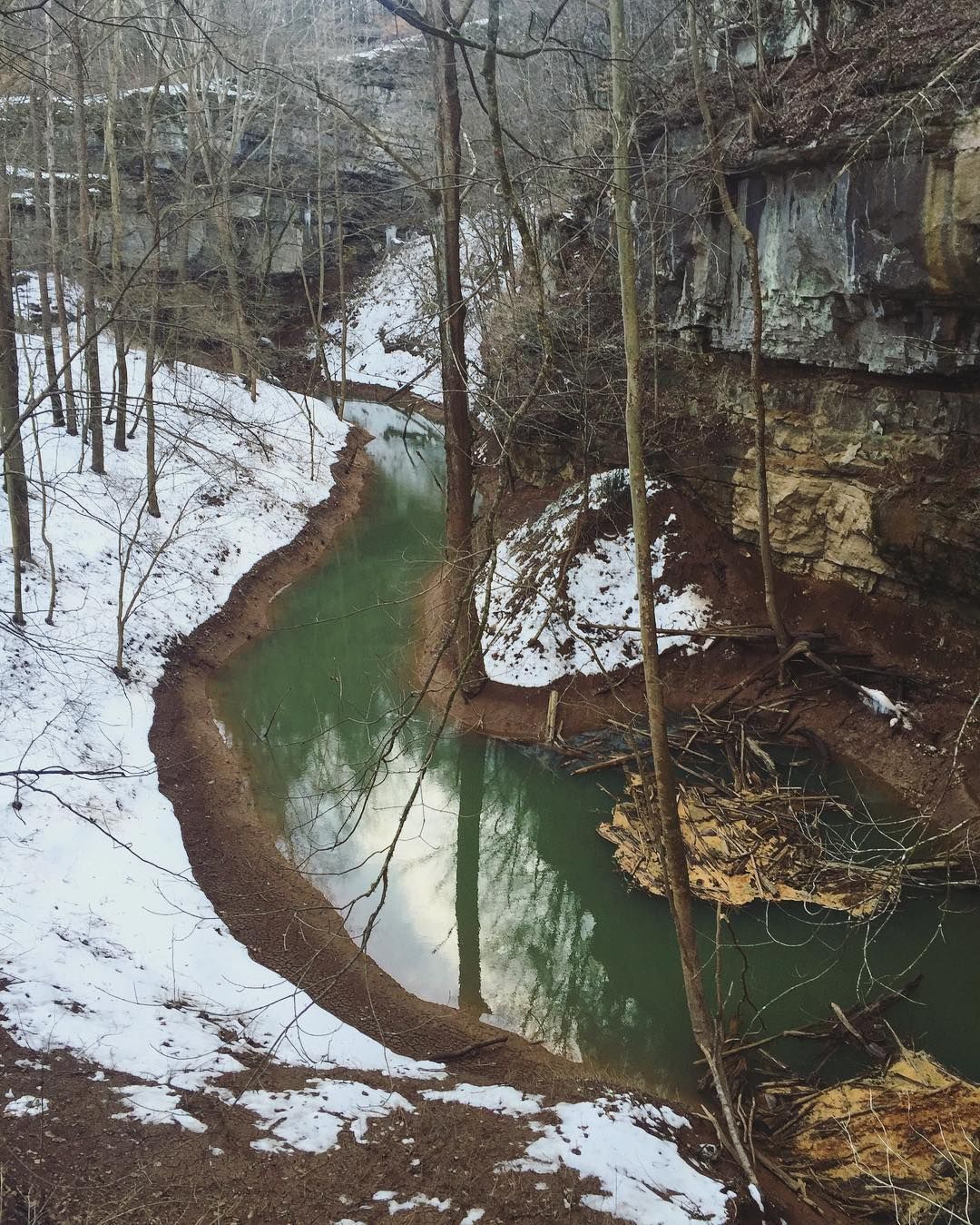 A winding green creek flows through a snow-dusted forest gorge with limestone cliffs in winter.