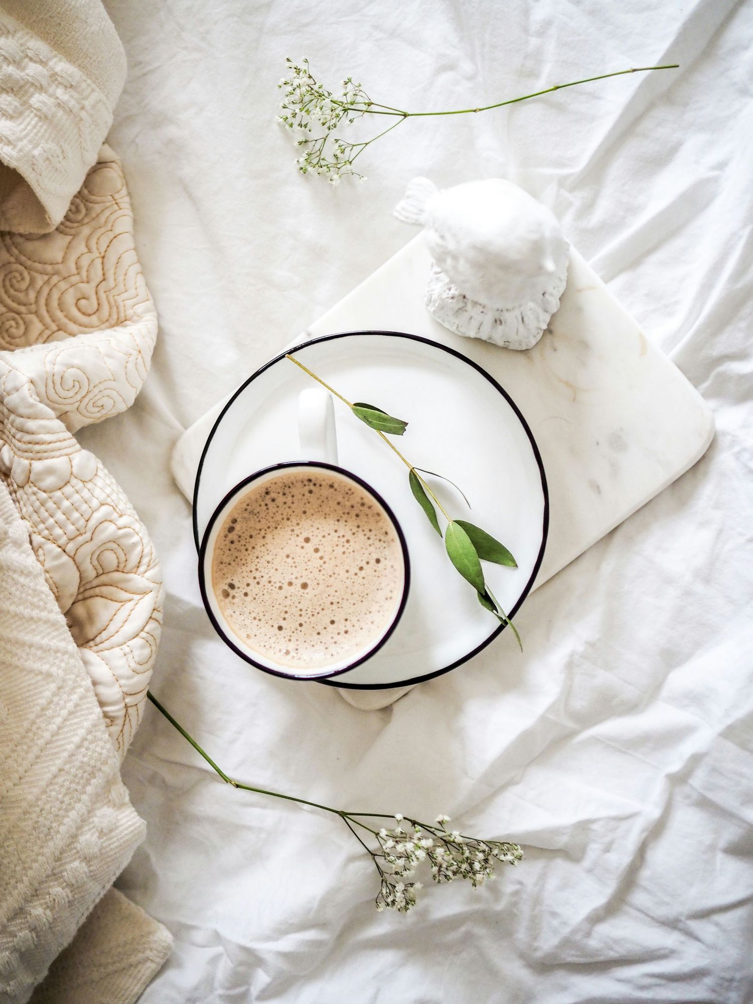A cozy flat lay of a frothy latte on a marble tray with eucalyptus and baby's breath on white linen bedding.