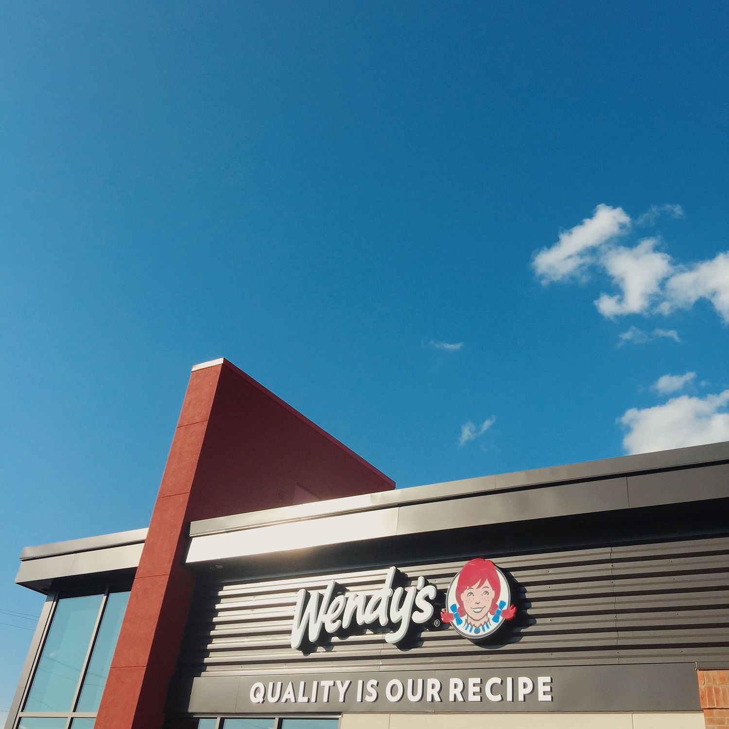 Wendy's fast food restaurant exterior with 'Quality Is Our Recipe' slogan under a bright blue sky with clouds.