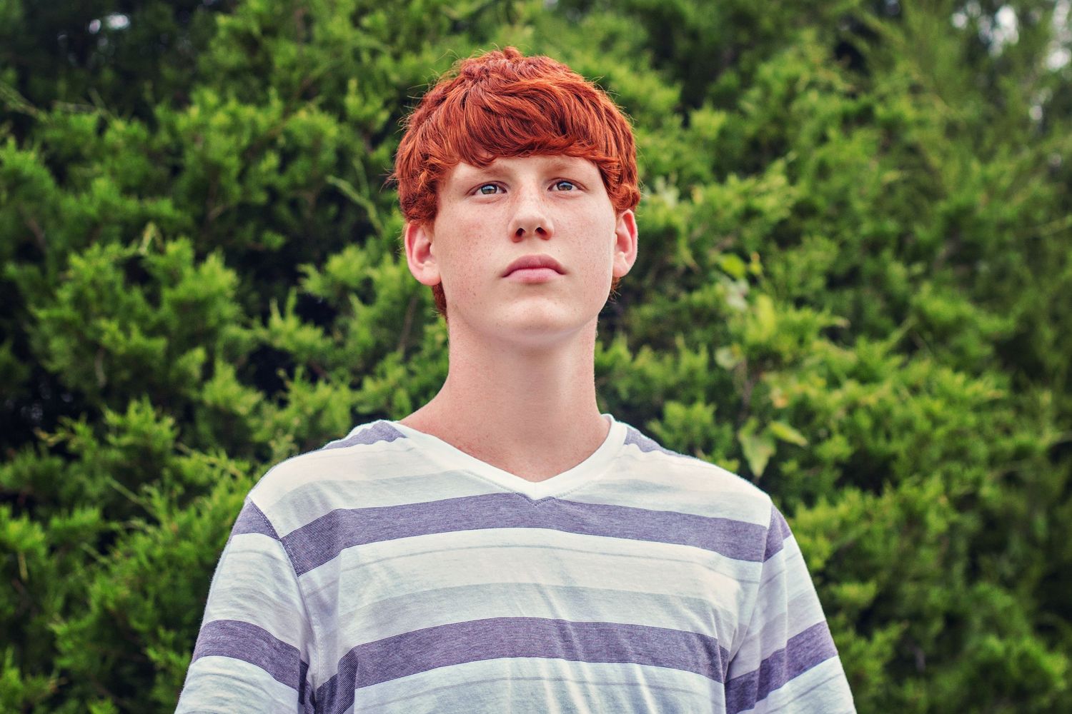 A young redhead teen boy in a striped shirt gazes upward outdoors with green trees in the background.