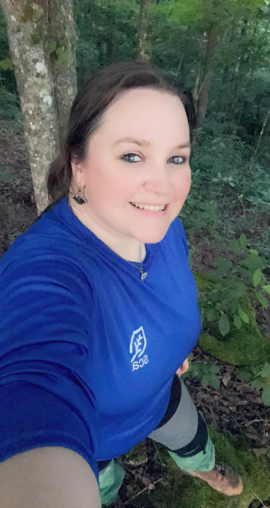 Selfie taken in a forest setting showing someone wearing a royal blue sweatshirt with a small logo emblem.