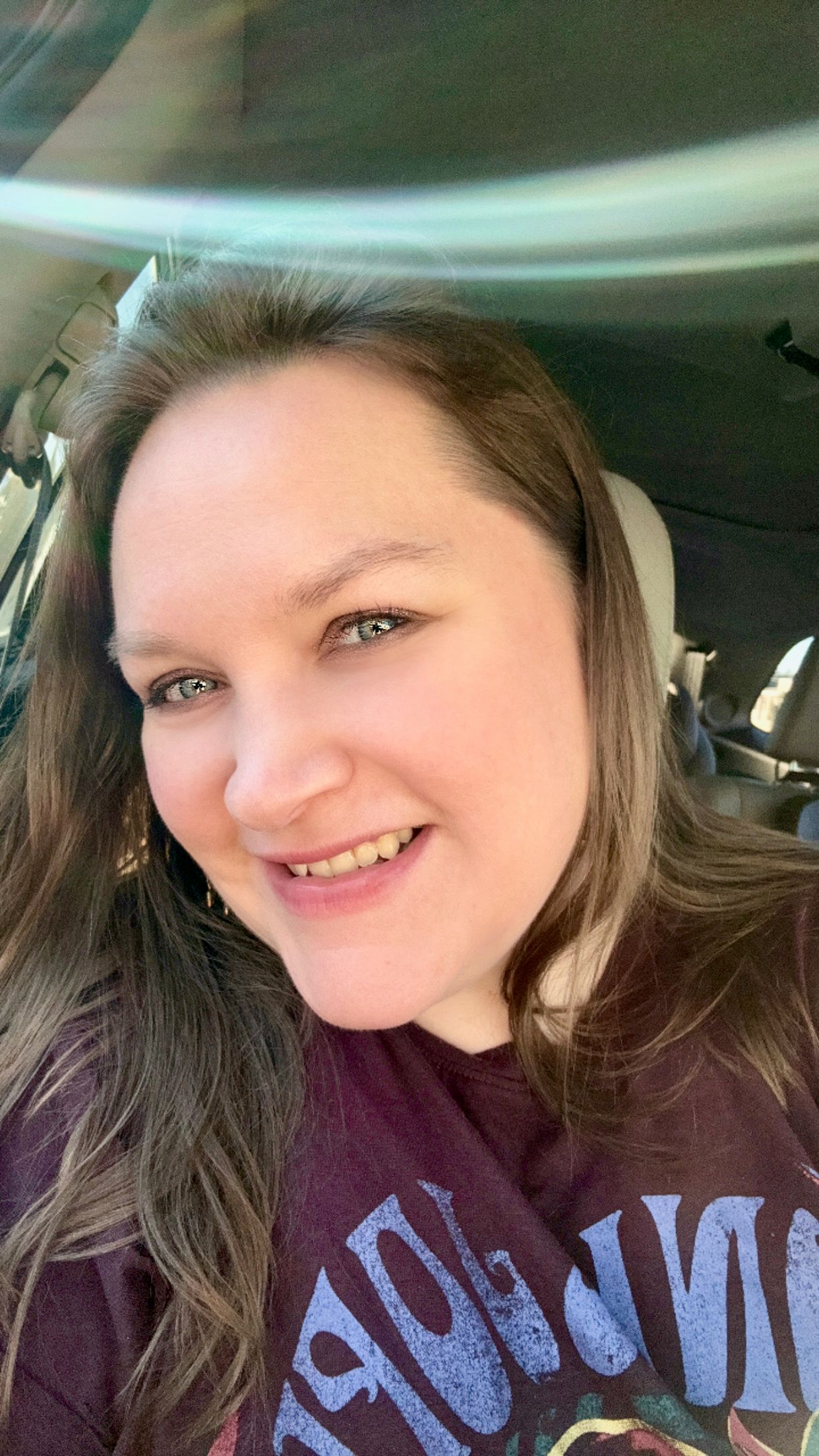 A smiling woman with long brown hair takes a selfie in a car, wearing a dark purple graphic t-shirt.