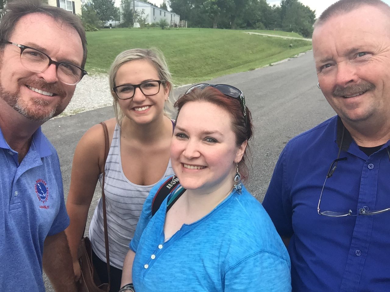 Four smiling adults posing for an outdoor selfie on a cloudy day near a green lawn and residential street.
