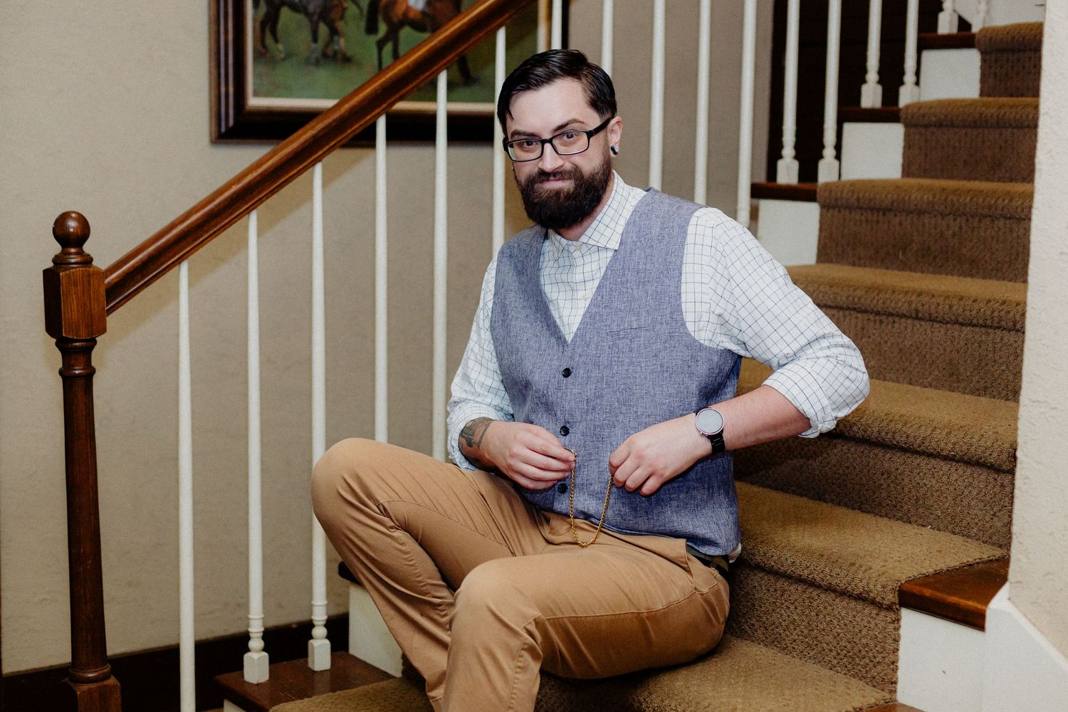 Bearded man in glasses and gray vest sits on carpeted staircase, smiling confidently in smart casual attire.