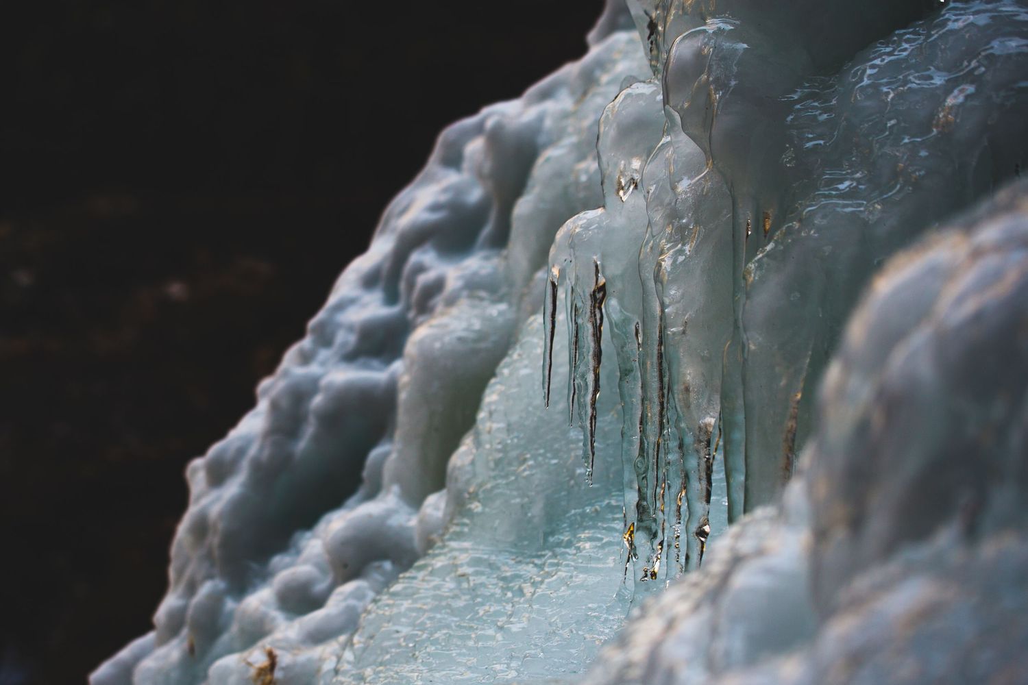 Close-up of a frozen waterfall with icicles and layered blue ice formations against a dark rocky background.