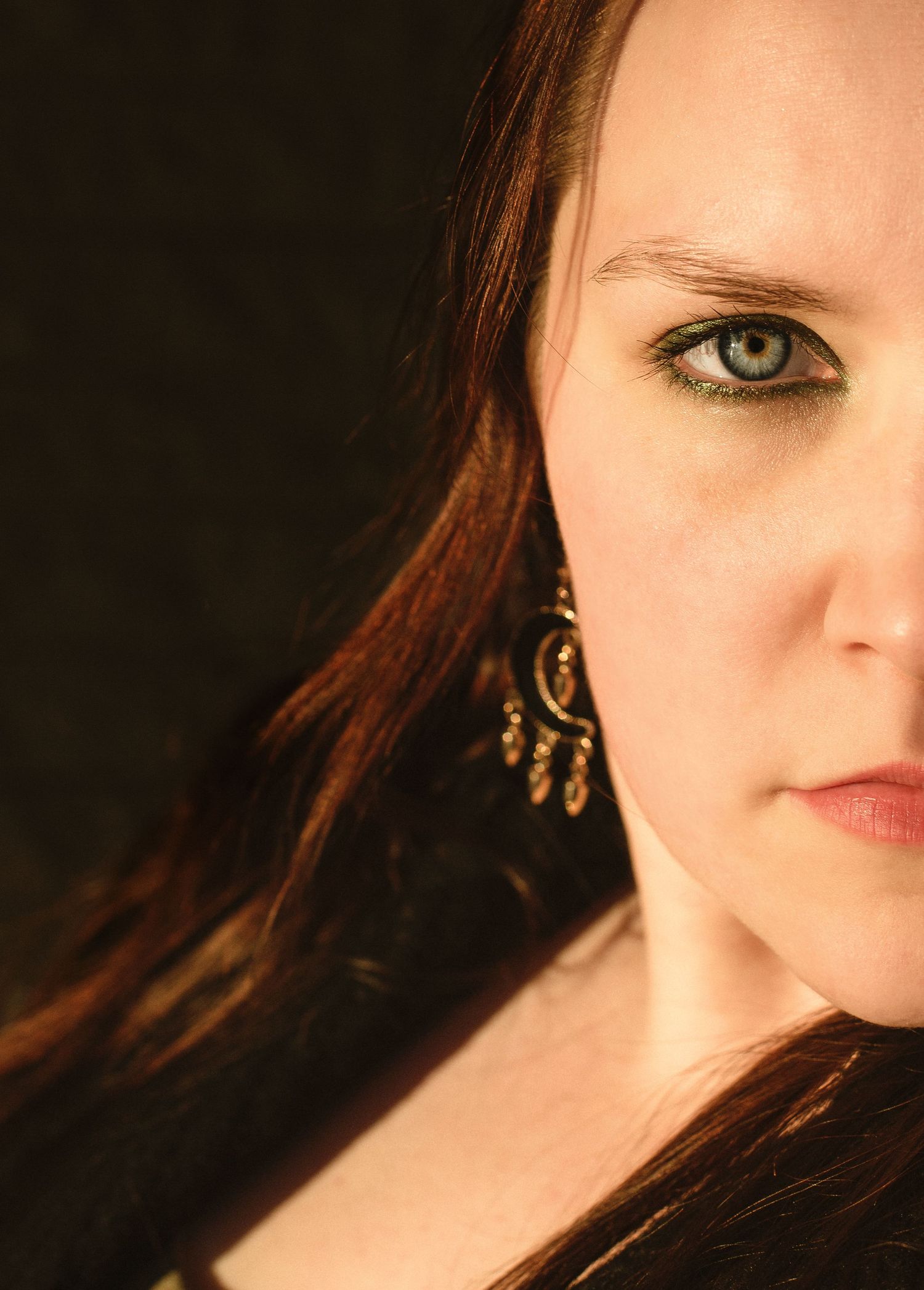 Close-up portrait of a woman with green eyes, red hair, and ornate gold earring against dark background.