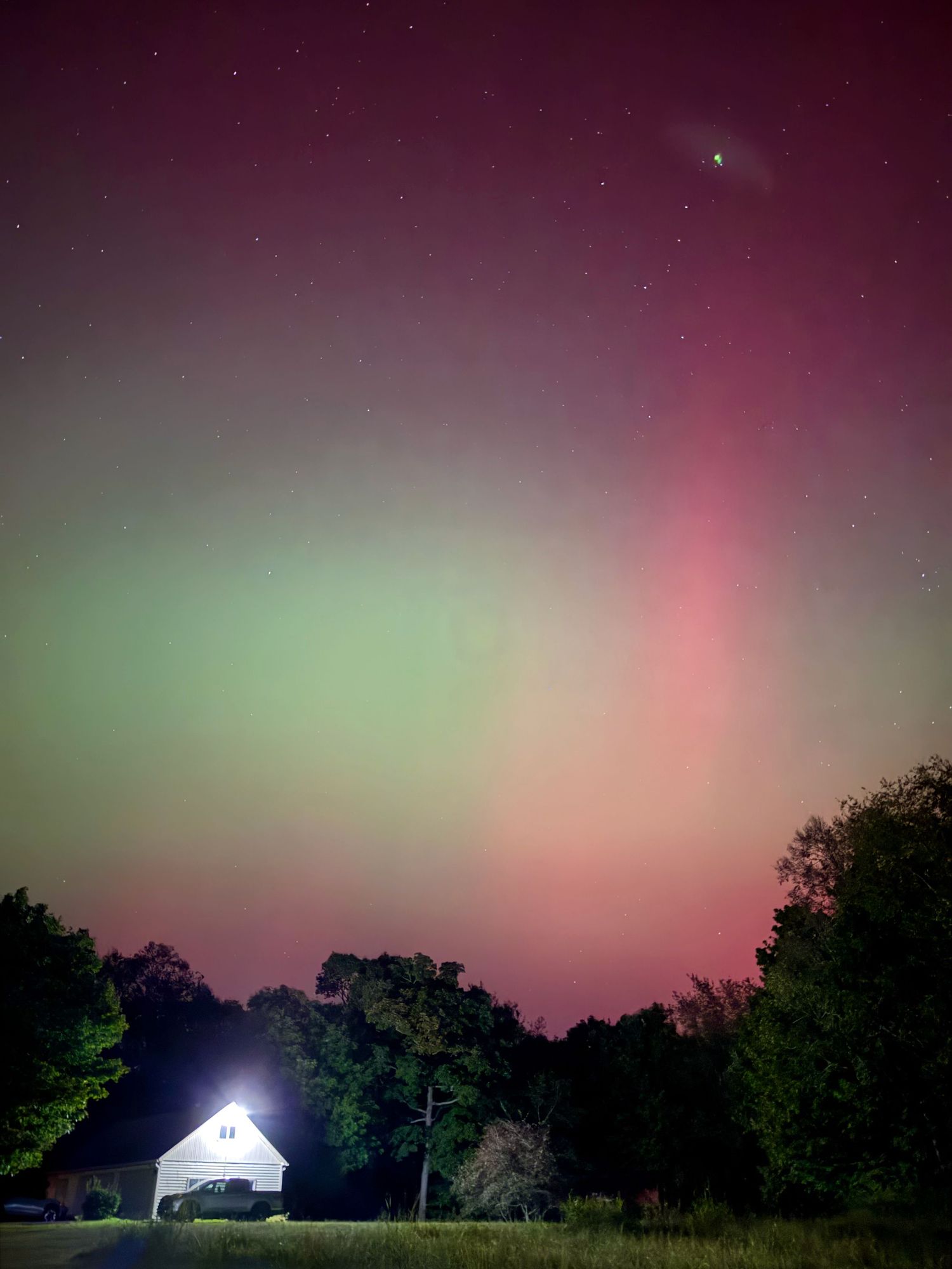 Aurora borealis displays green and pink colors over rural barn beneath starry night sky.