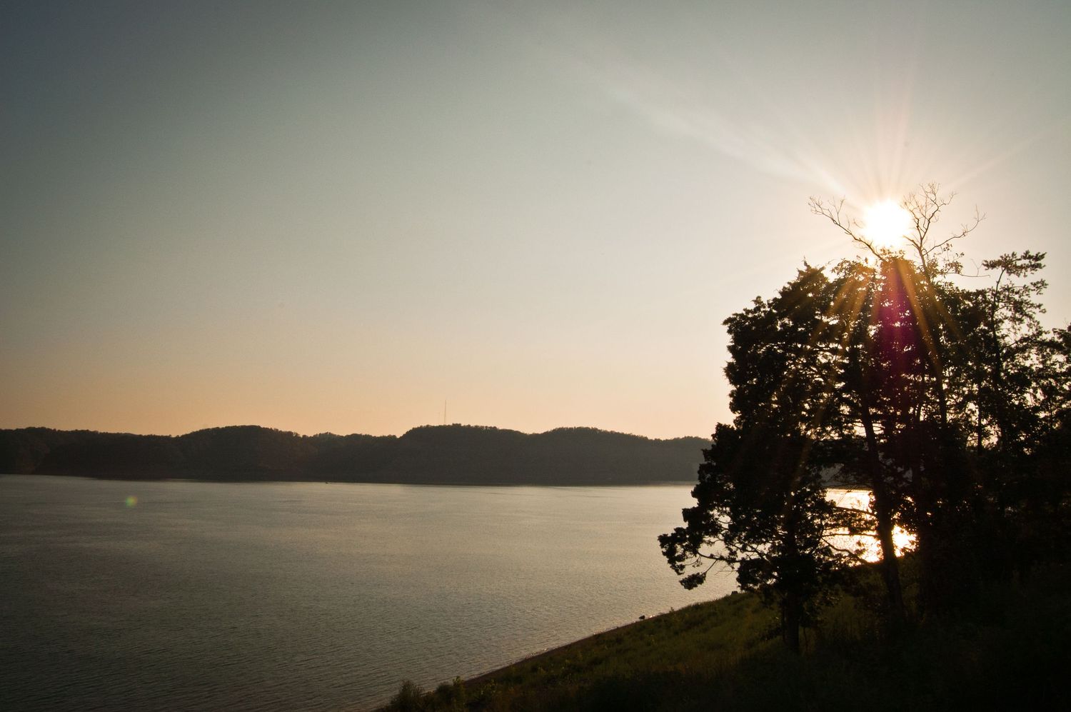 Sunset over a calm lake with silhouetted tree and rolling hills in the background.