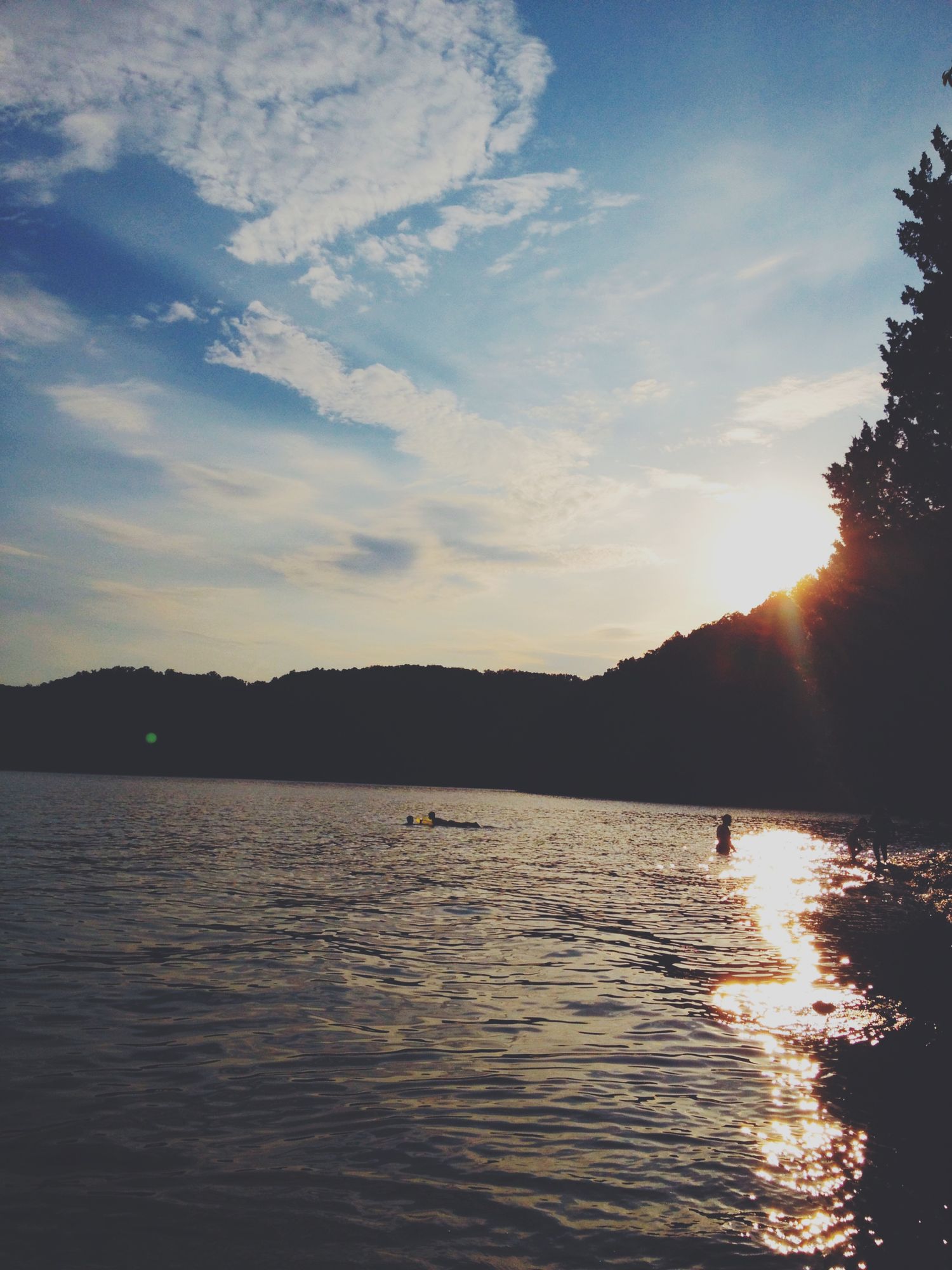 A scenic sunset view of swimmers in a lake surrounded by silhouetted trees and hills with sunlight sparkling on the water.