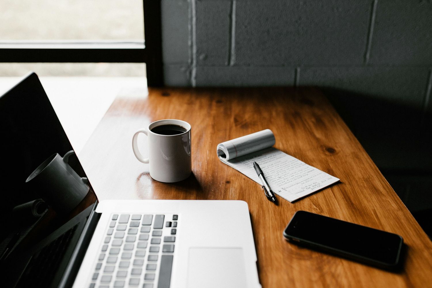 Laptop, coffee mug, notepad, pen, and smartphone on a wooden desk near a window in a modern workspace.