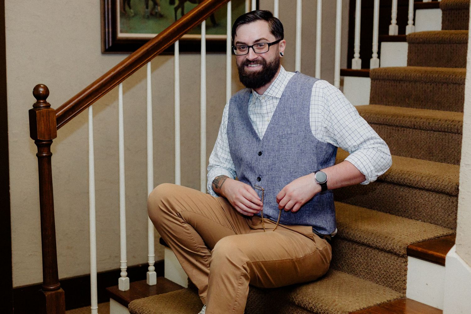 A person in a gray vest and khaki pants sits casually on carpeted stairs near a wooden handrail.