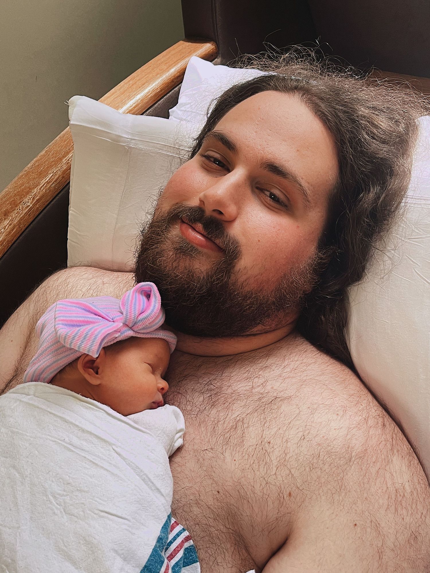 A bearded man with long hair lies in a hospital bed doing skin-to-skin contact with a sleeping newborn baby girl.